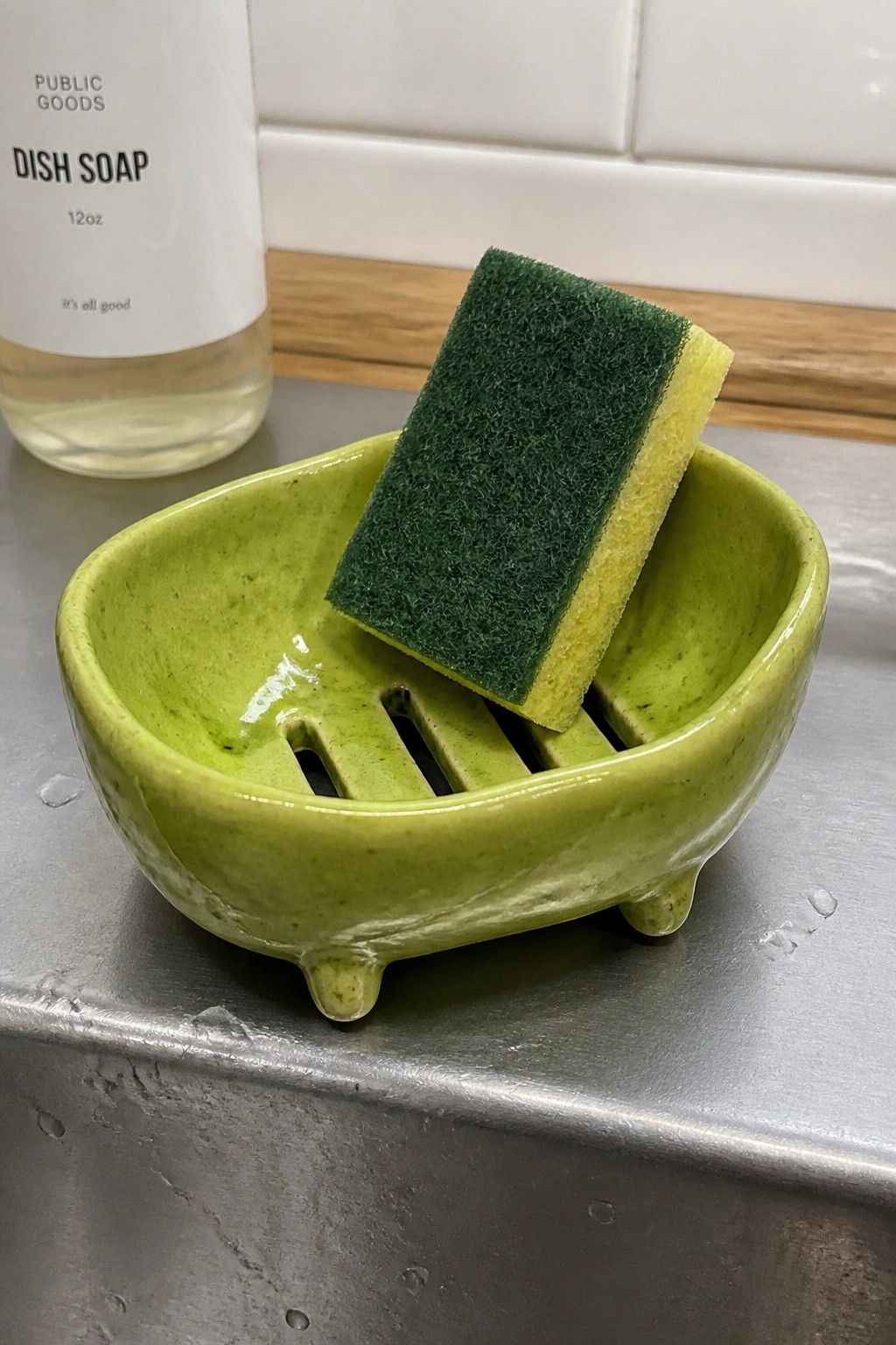 Green ceramic dish with green-yellow sponge on stainless steel sink edge.