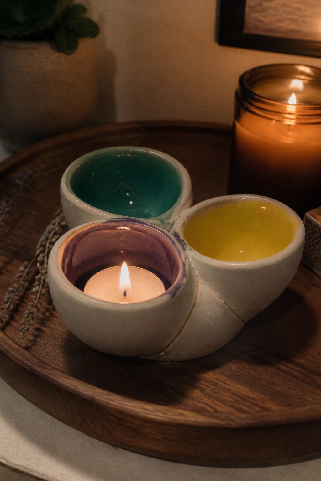 A handmade ceramic holder with three connected bowls in teal, yellow, and purple glazes sits on a wooden tray, one bowl holding a lit tea light candle.