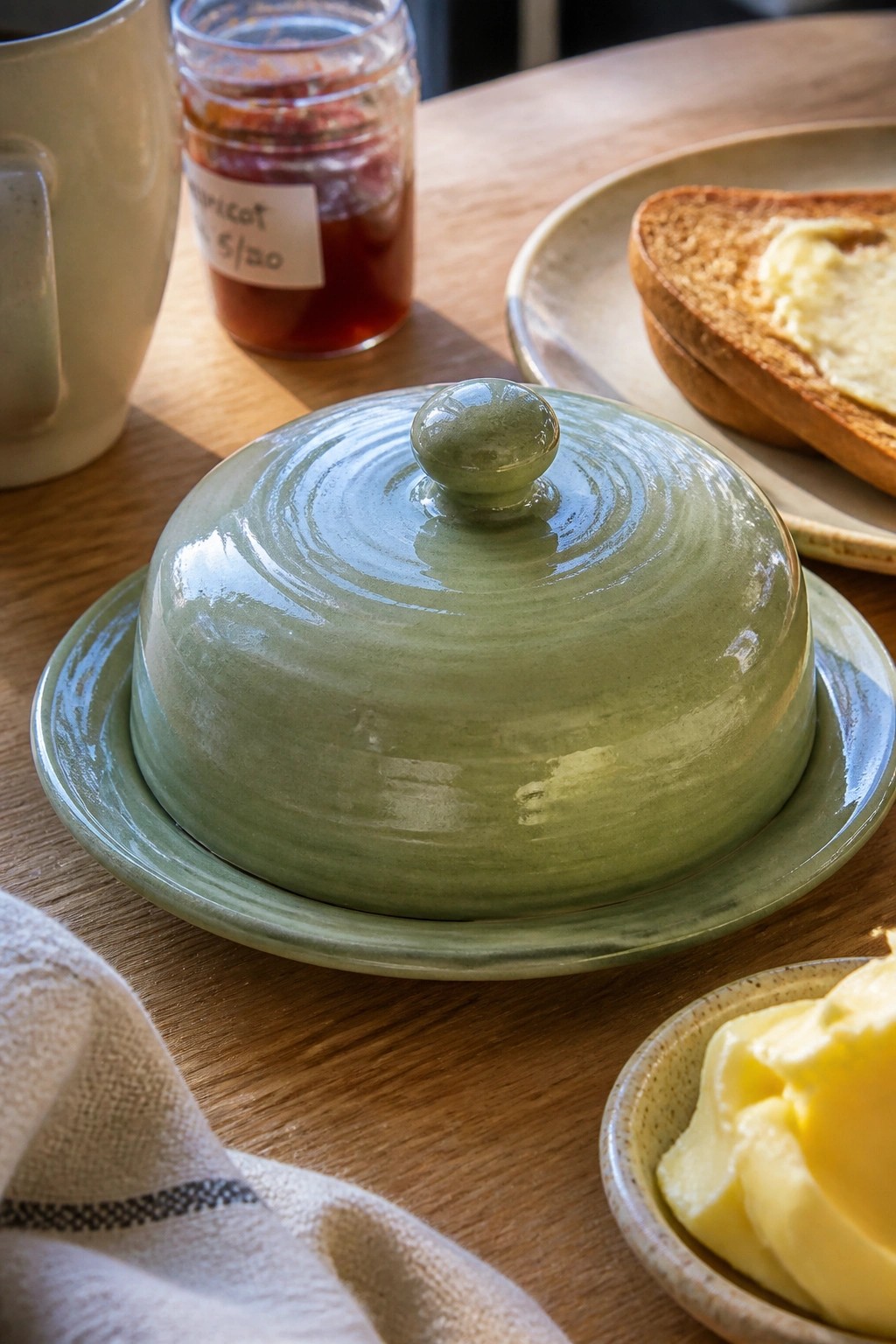 Green ceramic butter dish on wooden table with toast, jam, and butter