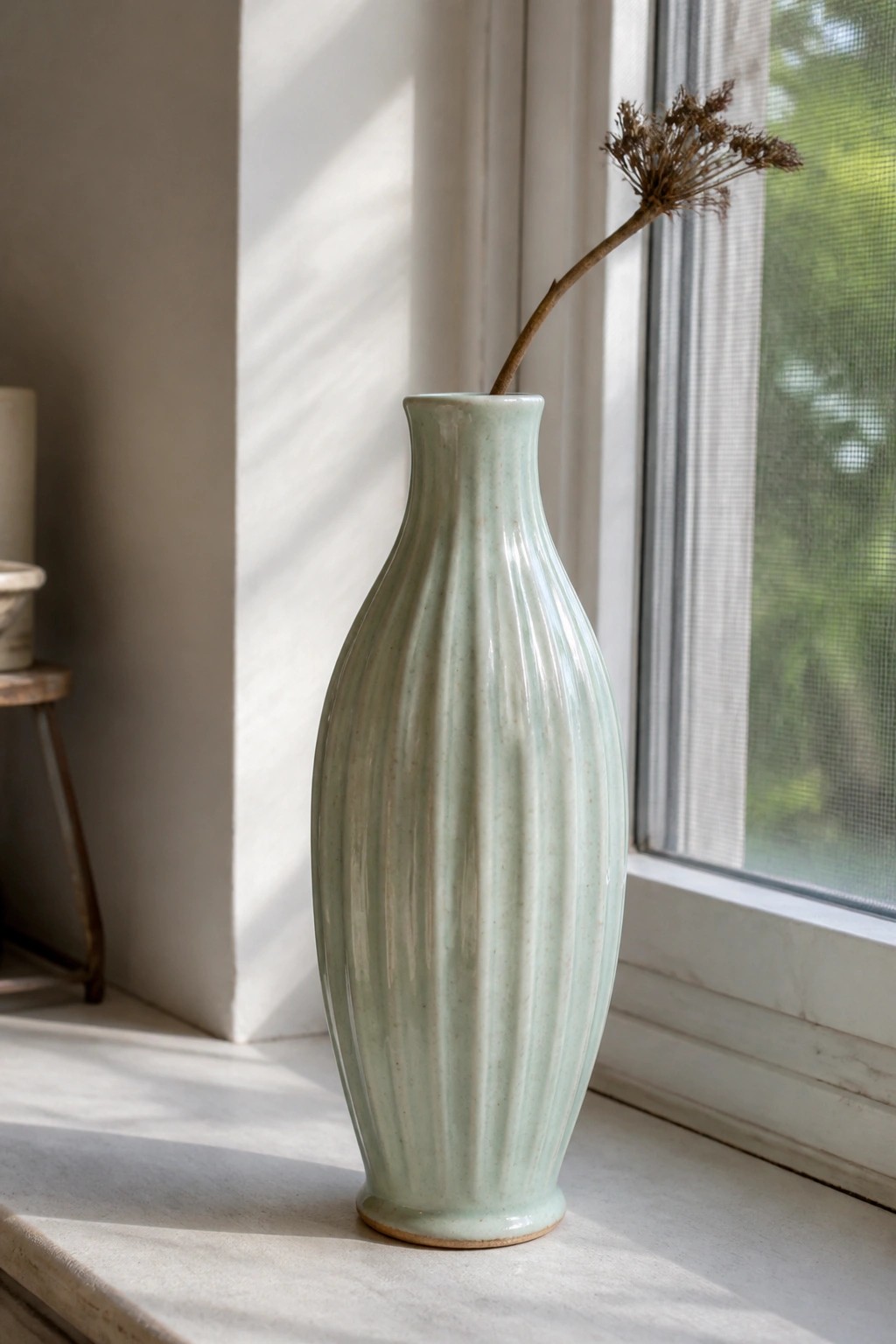A light green ceramic vase with vertical ridges sits on a windowsill holding a dried flower stem.
