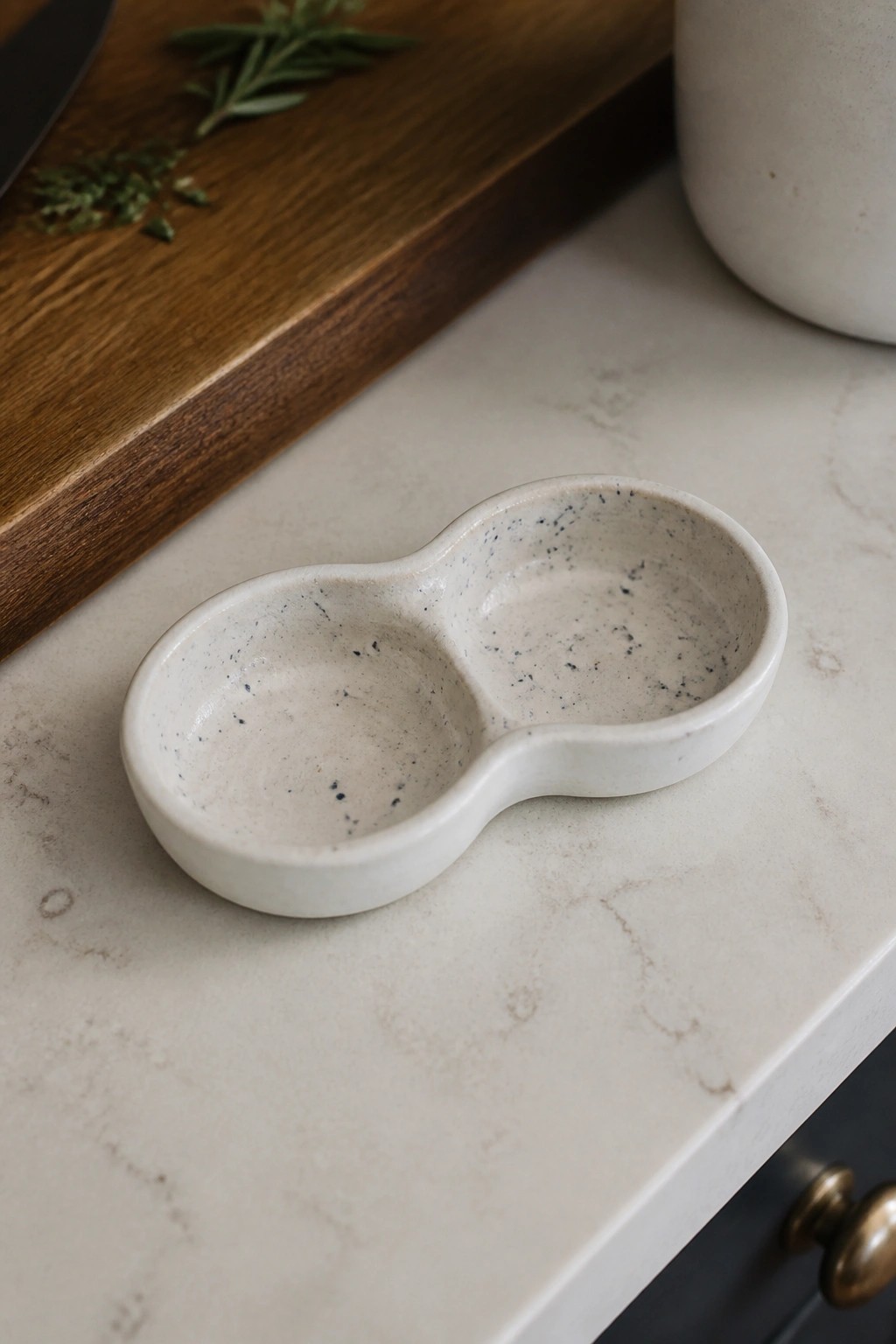Speckled white ceramic two-compartment bowl on marble countertop with wooden board and herbs