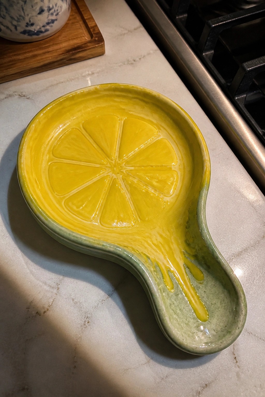 A ceramic lemon-shaped spoon rest with yellow glaze dripping down the handle sits on a marble countertop.