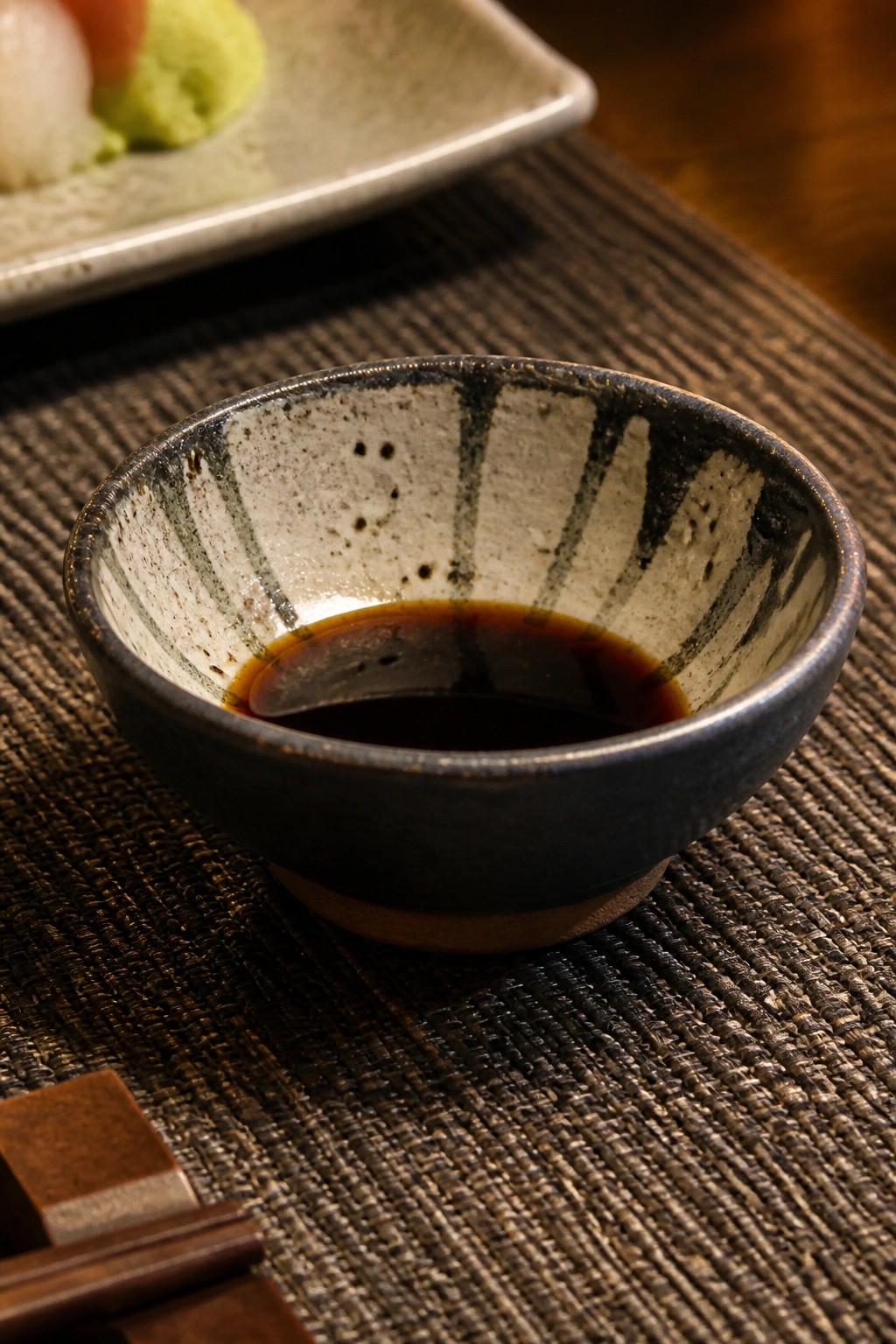 Speckled ceramic bowl of soy sauce on woven mat beside sushi and chopsticks