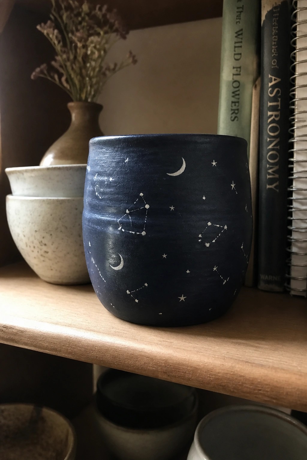 A dark blue ceramic mug with white painted stars, moons, and constellations rests on a wooden shelf beside books and other pottery.