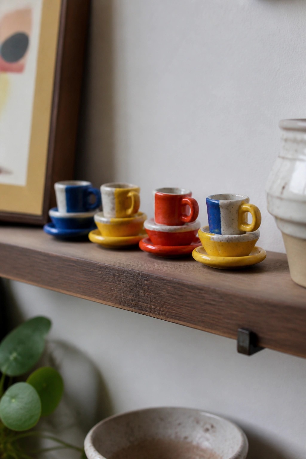 Small colorful ceramic cups and saucers in blue, yellow, and red sit on a wooden shelf.
