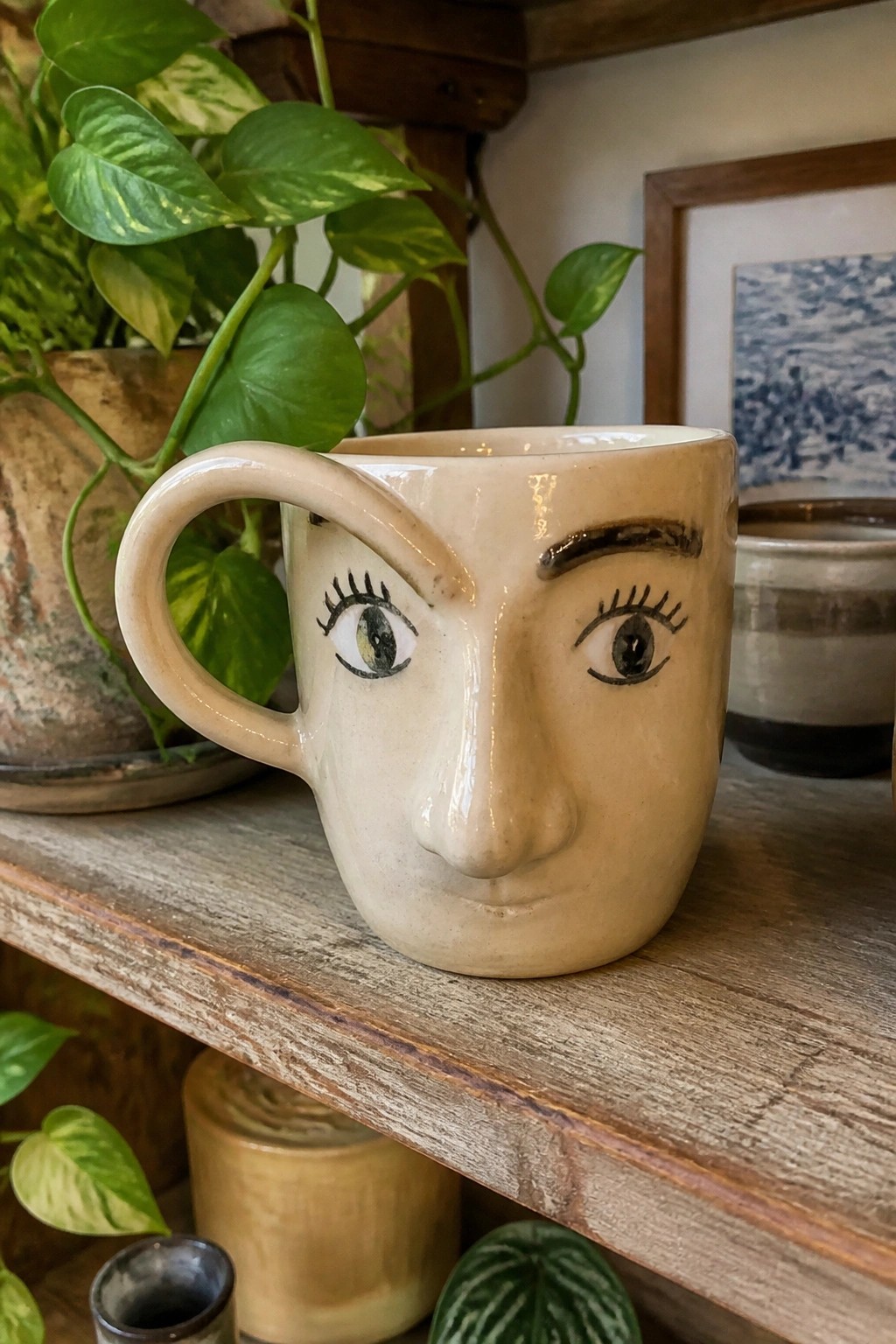 Beige ceramic face mug with painted eyes on wooden shelf among plants