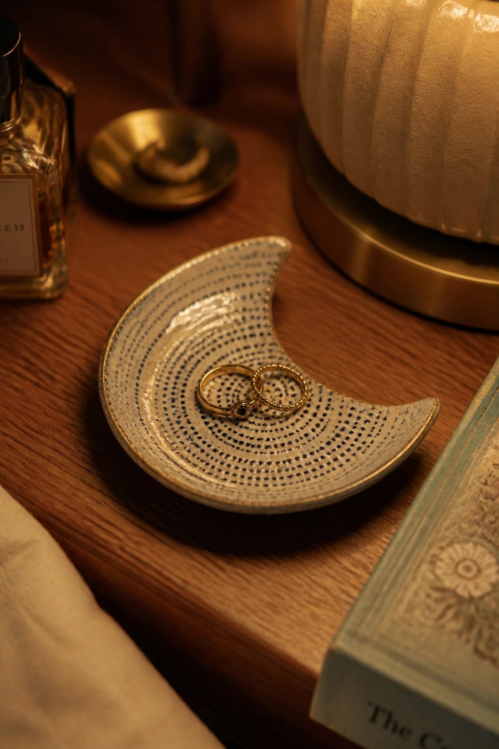 A crescent-shaped ceramic ring dish with a dark dotted pattern and gold rim holding two gold rings on a wooden surface.
