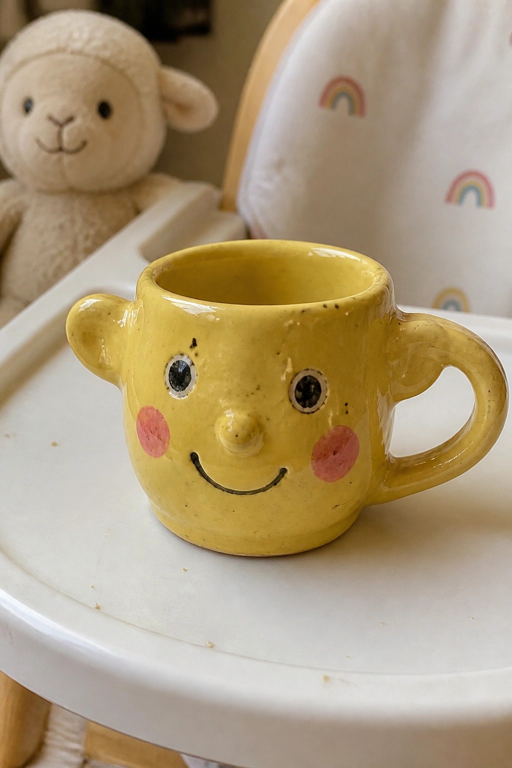 Yellow ceramic face mug on white high chair tray beside stuffed lamb