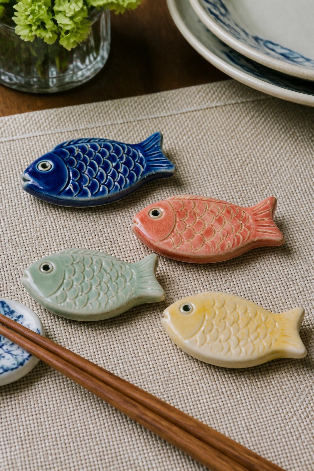 Four colorful ceramic fish on woven mat beside wooden chopsticks and blue plates.