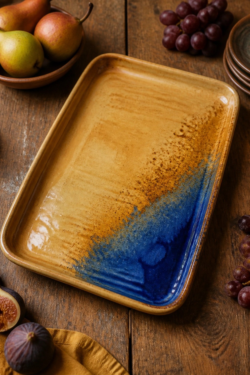 Rectangular ceramic tray with blue-brown glaze amid pears, grapes, and figs on wood