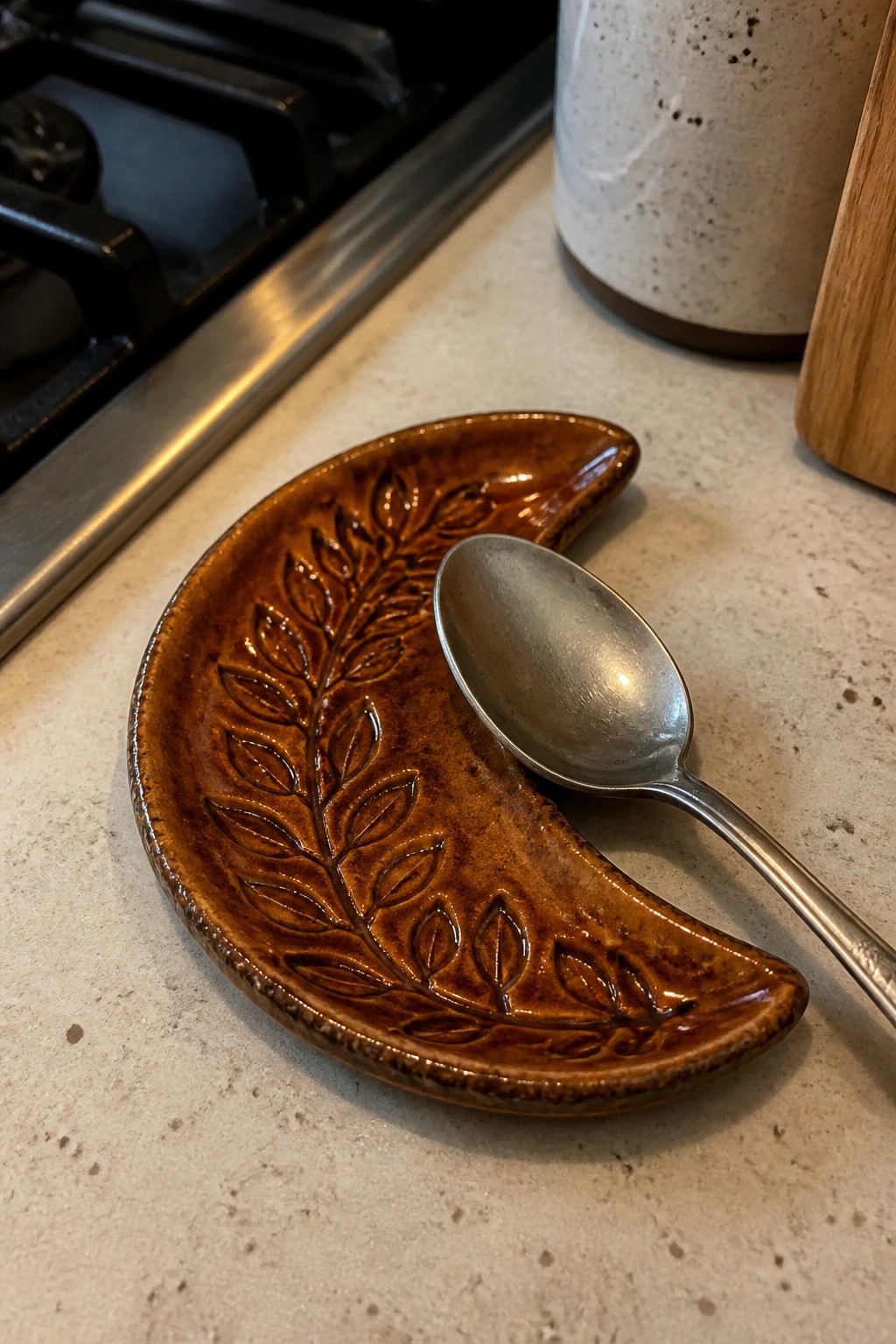 Brown ceramic leaf-embossed spoon rest holding a metal spoon on kitchen counter
