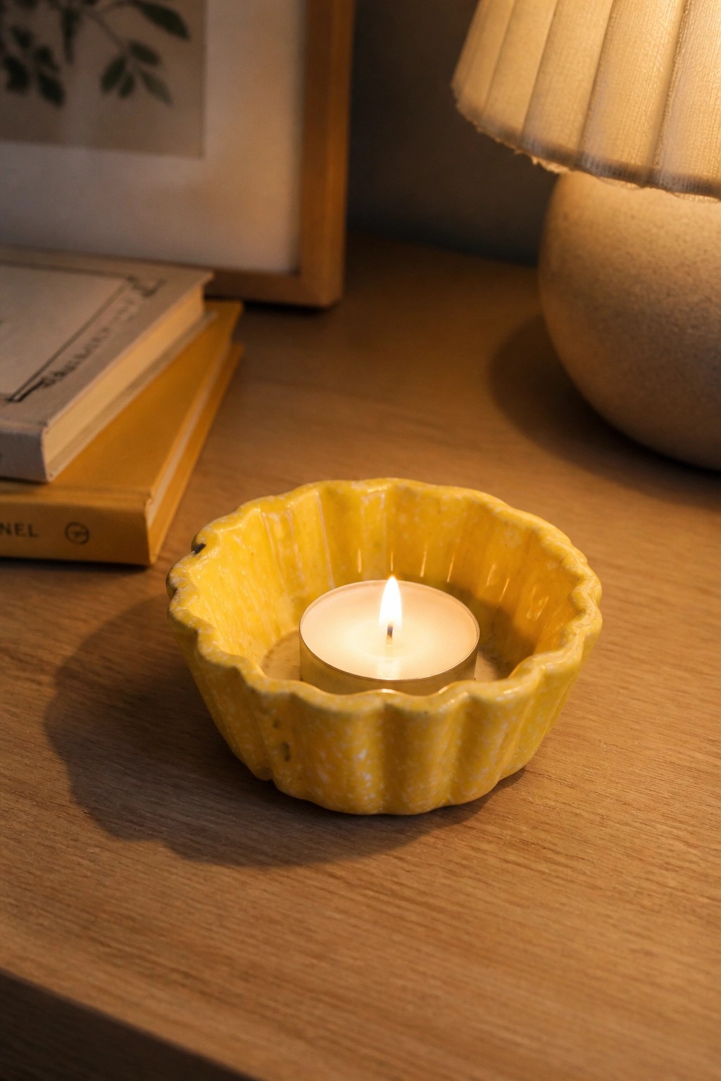 Lit candle in yellow ceramic holder on wooden table with books and lamp