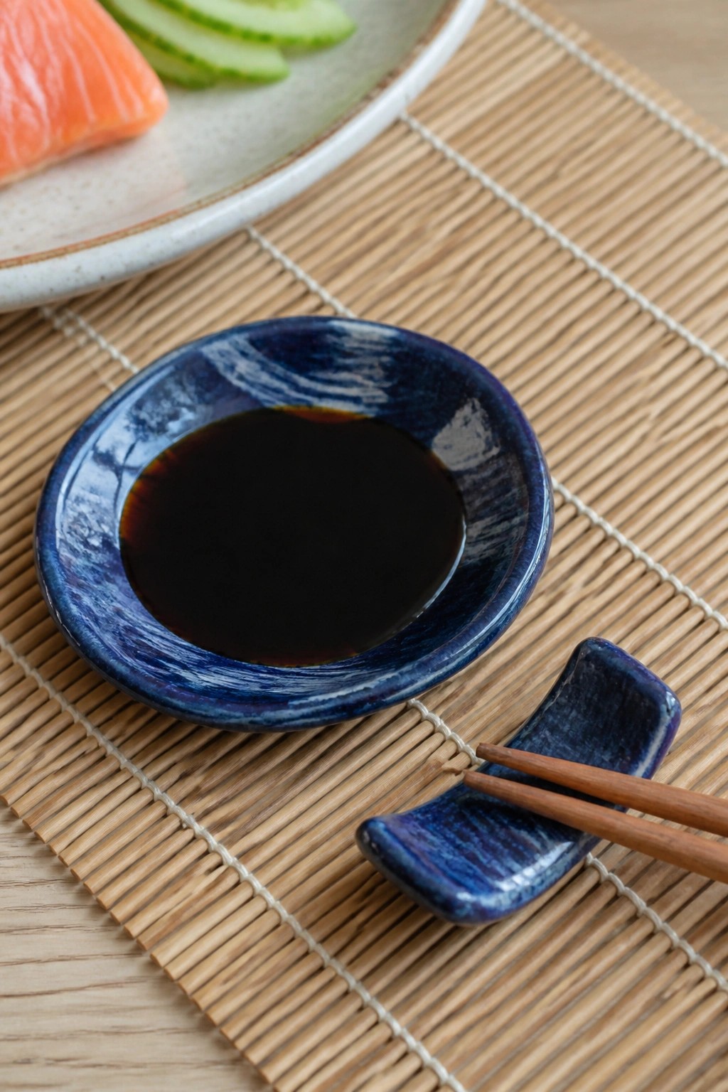 Blue soy sauce bowl with chopsticks on bamboo mat near salmon and cucumber