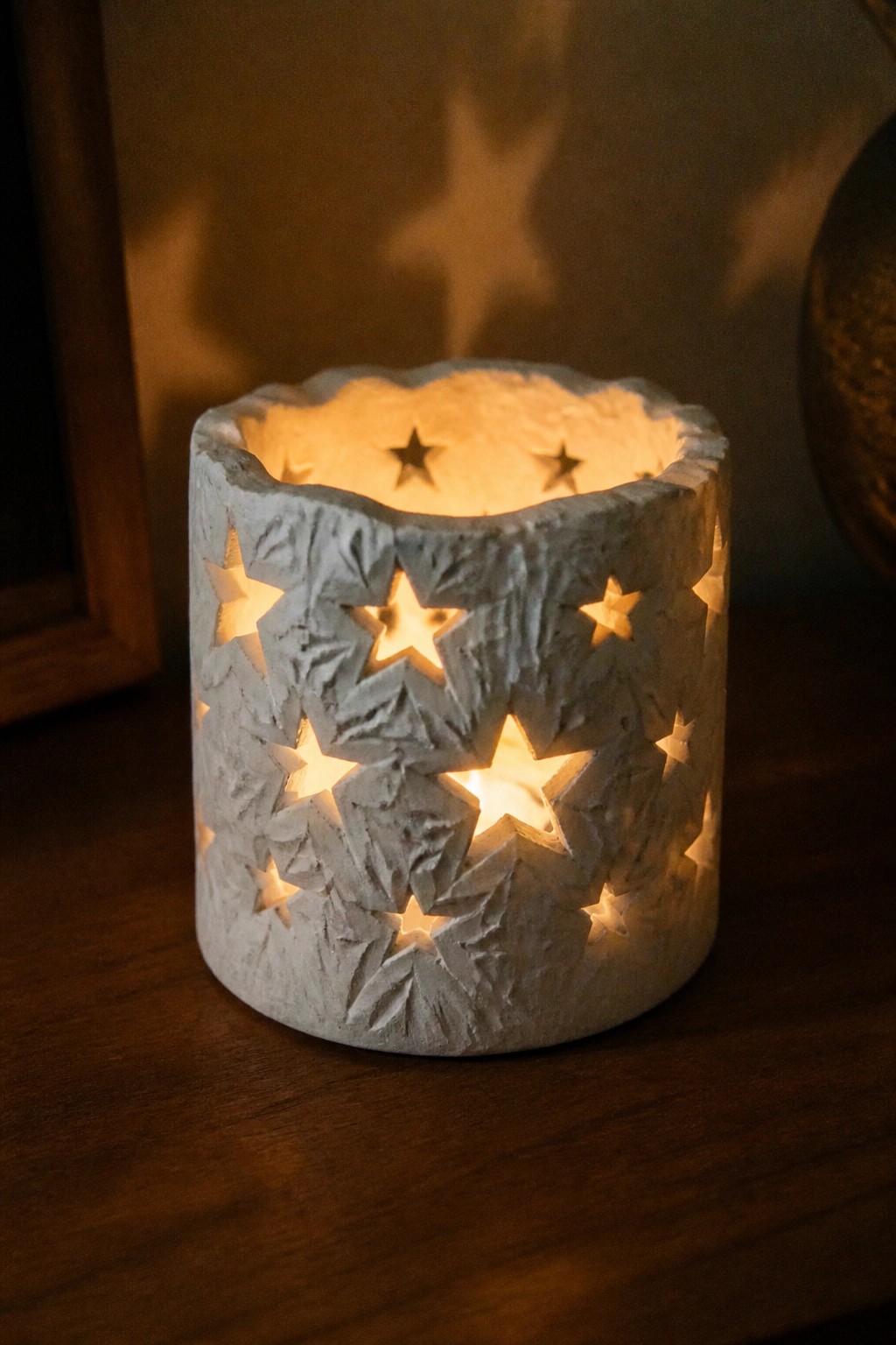 A white clay cylinder with multiple star cutouts glowing from an internal light on a wooden surface.