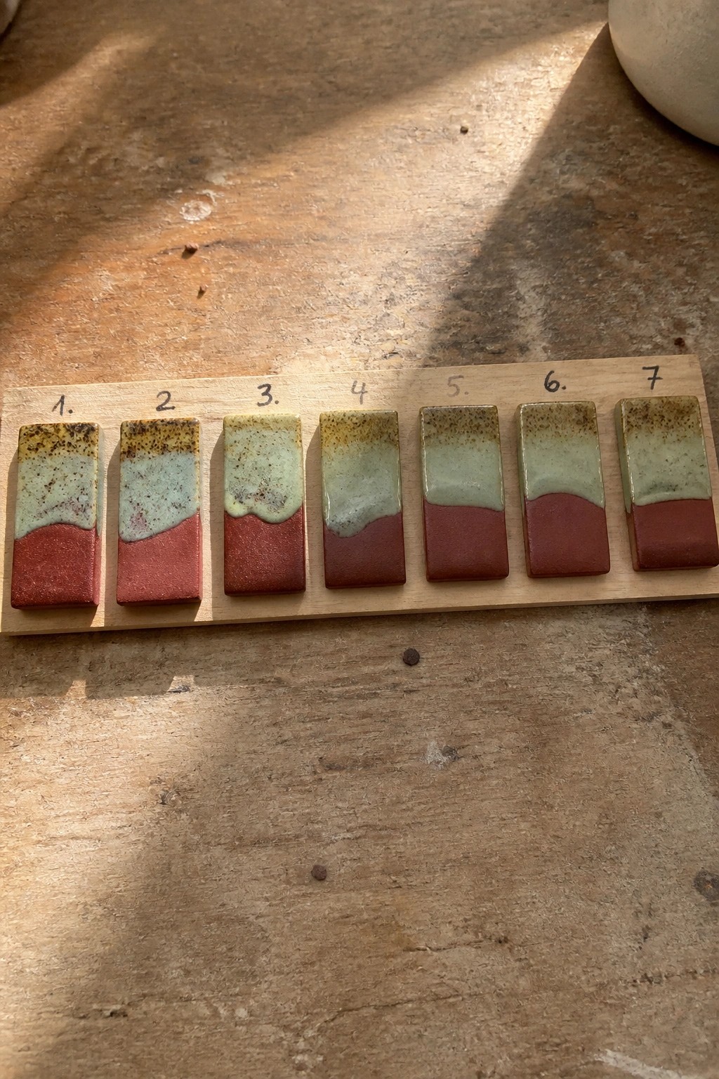 Seven numbered ceramic tiles on wood with red bases and speckled green glazes.