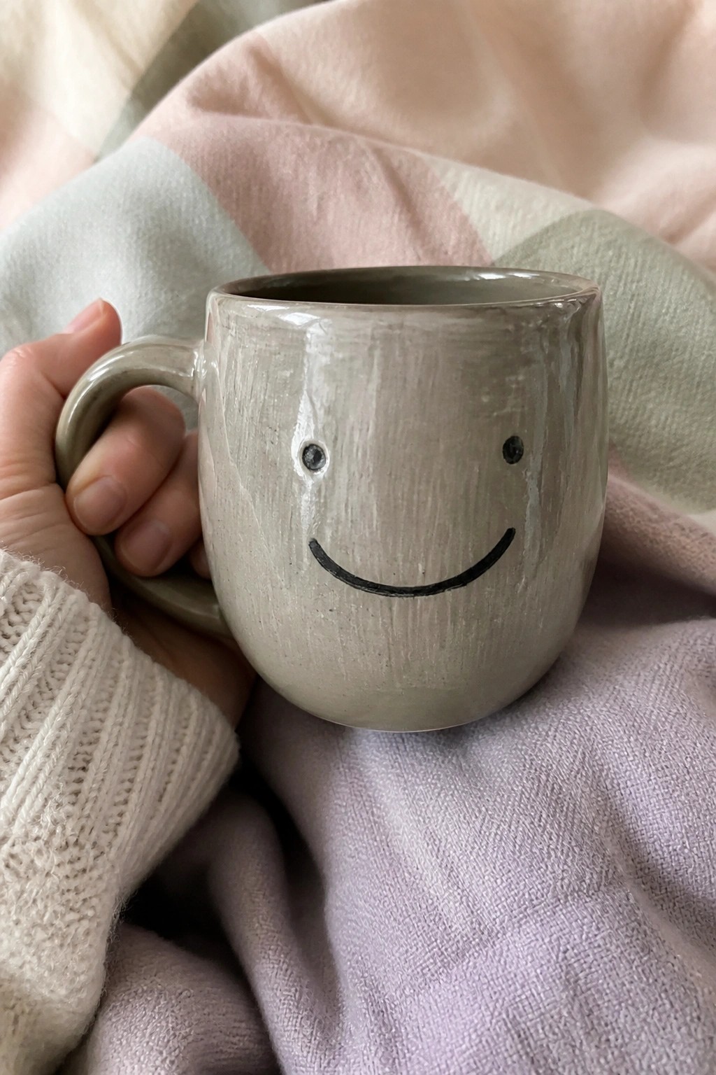 Hand in white knit sweater holding smiling gray ceramic mug on pastel plaid blanket.