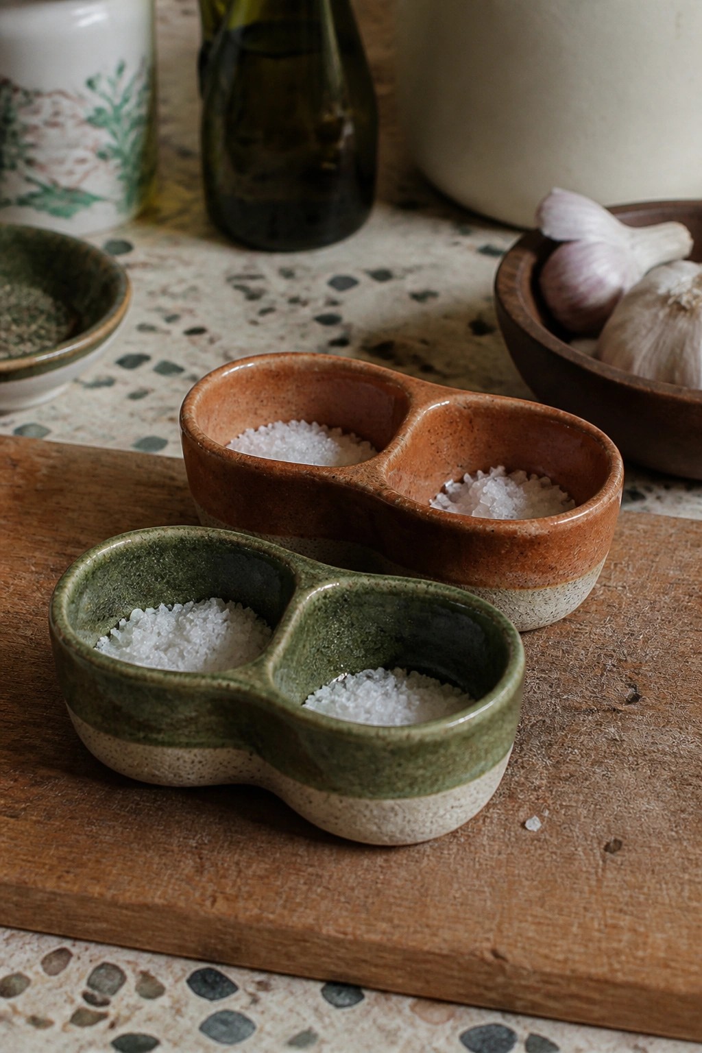 Two ceramic salt cellars on wooden board with garlic and bottles nearby.