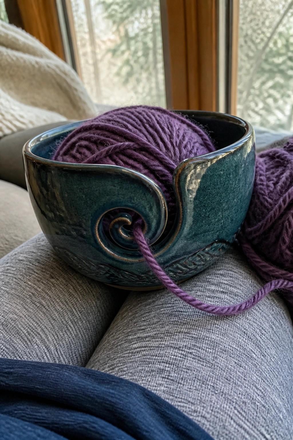 A teal ceramic yarn bowl holding purple yarn with the strand pulled through the side spiral opening, resting on gray fabric.