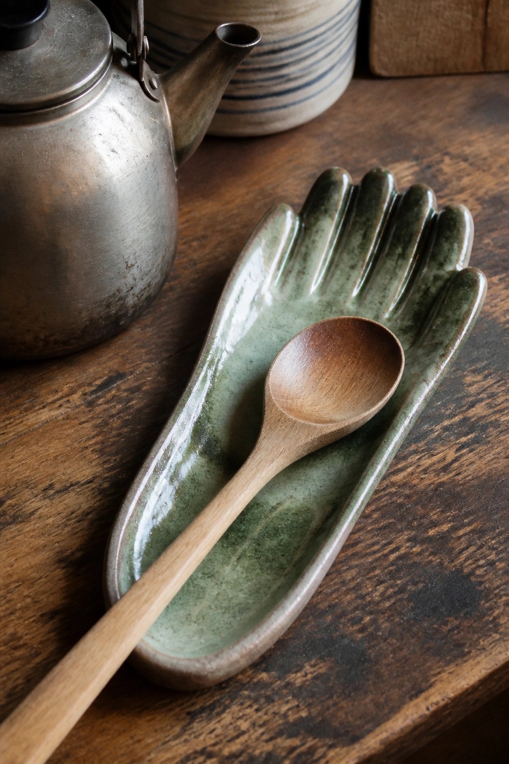 Wooden spoon on green ceramic hand-shaped dish beside metal teapot on wooden table