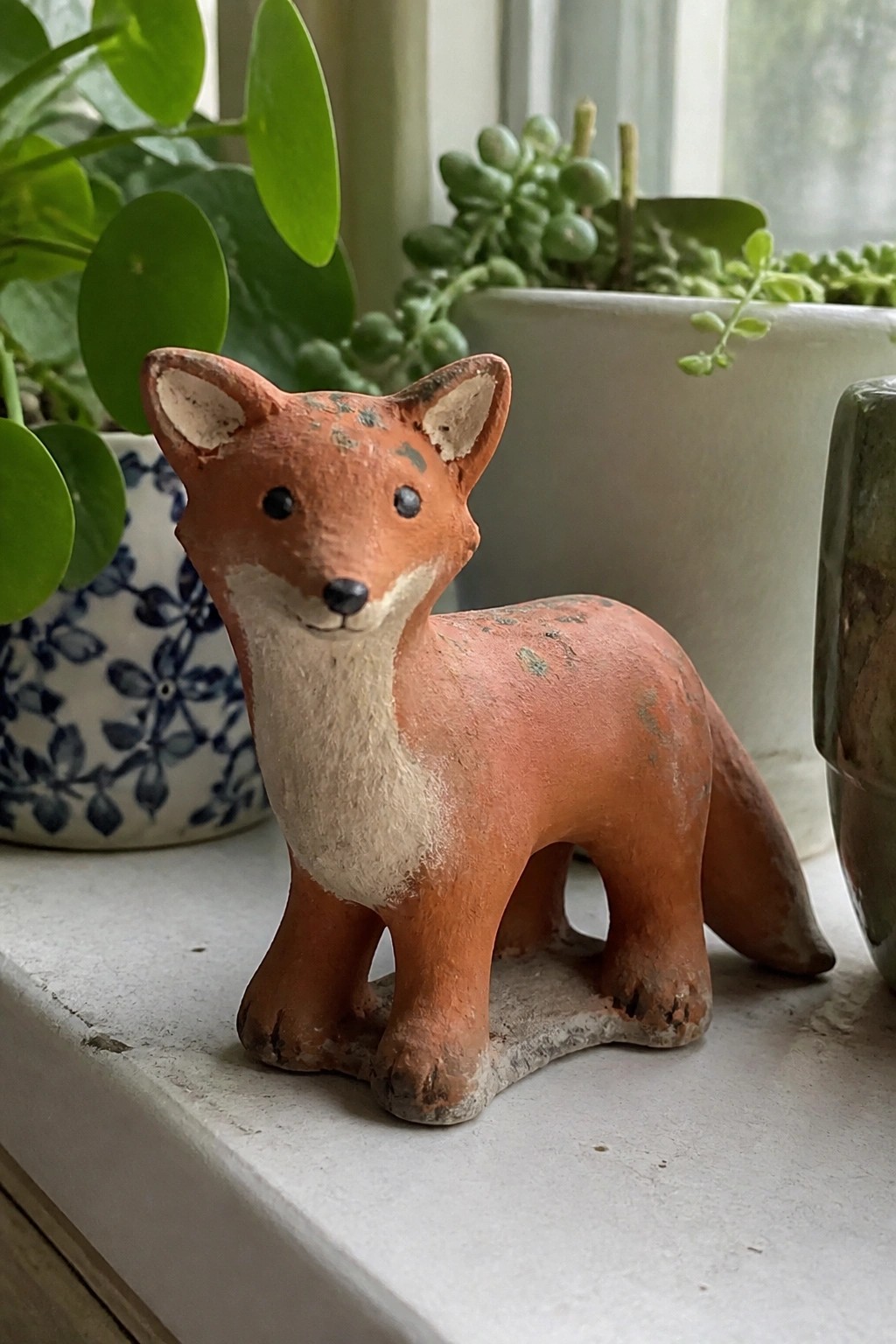 Rustic orange ceramic fox figurine on windowsill amid green potted plants