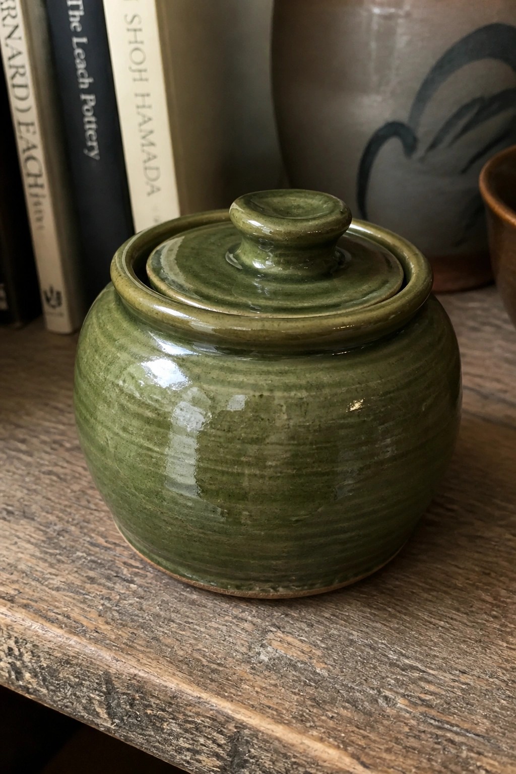 Green ceramic lidded jar on wooden table with books and pottery behind