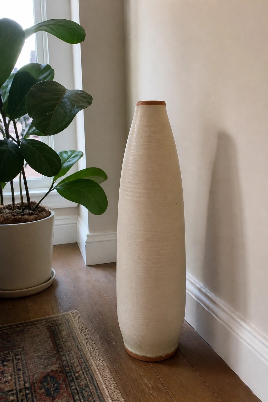 Tall off-white vase beside potted green plant on wooden floor
