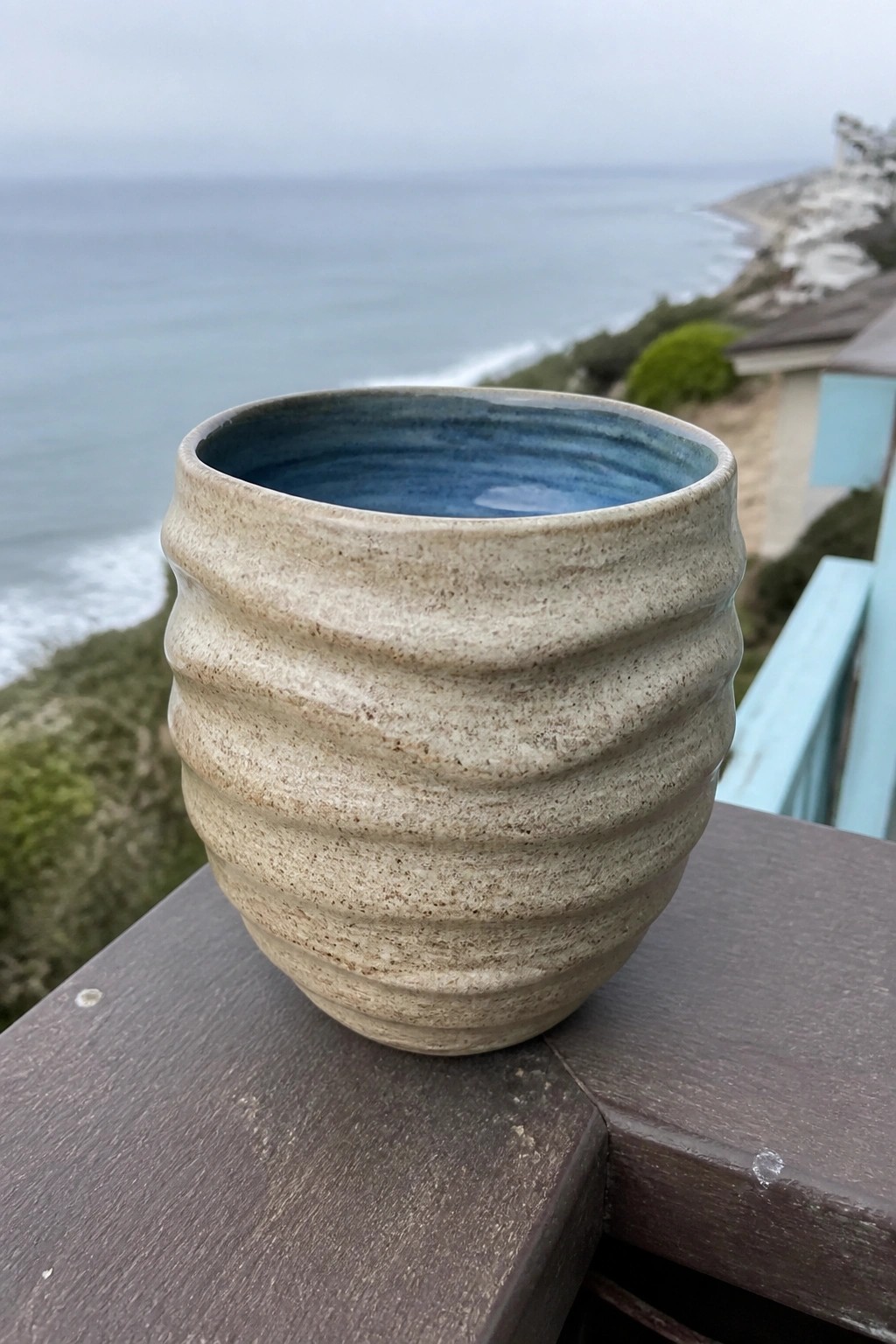Beige ridged ceramic cup with blue interior on wooden railing overlooking ocean