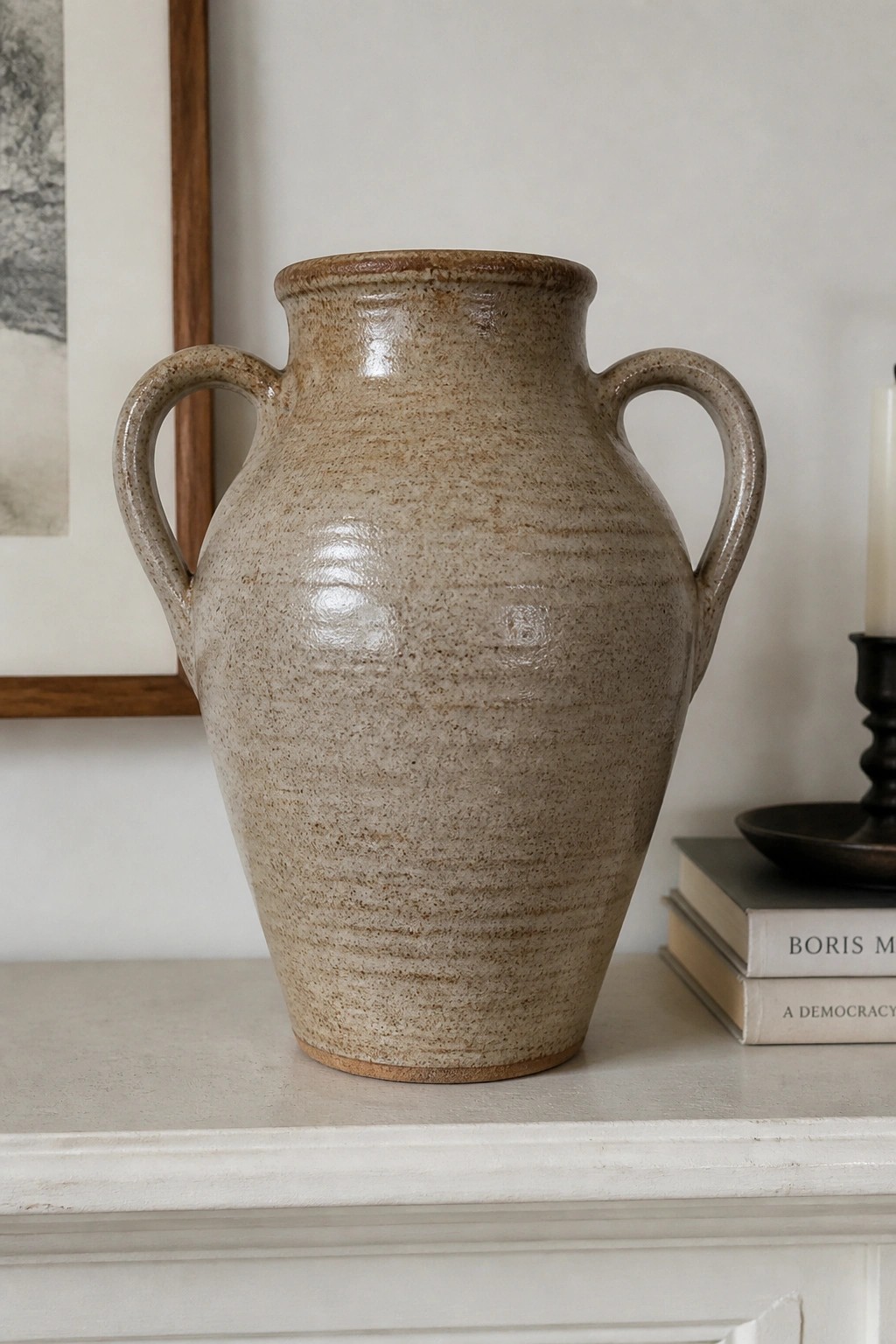 A large ceramic vase with two handles and a speckled beige glaze sits on a white mantel next to stacked books.