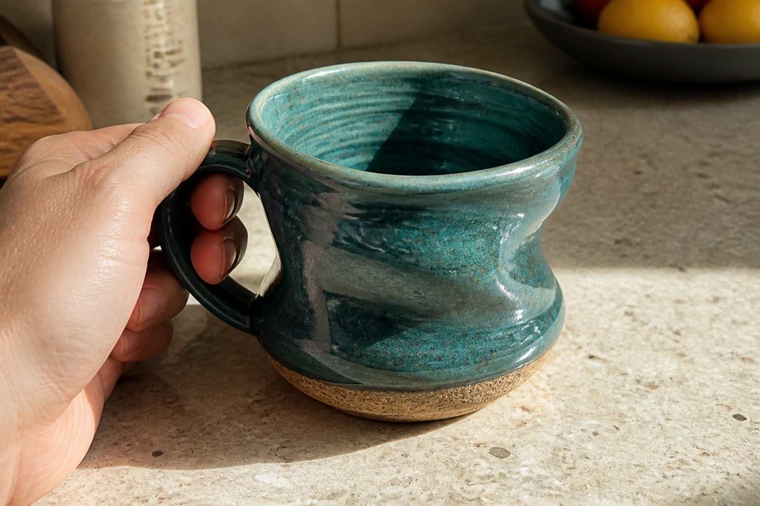 Hand holding a glossy teal ceramic mug with face design on sunny counter