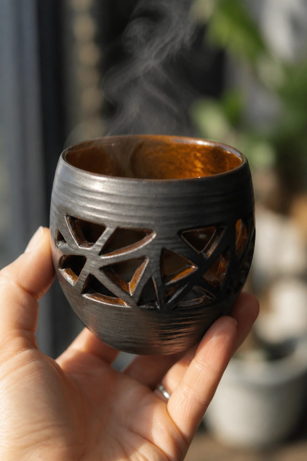 Hand holding steaming dark ceramic cup with intricate geometric cutouts