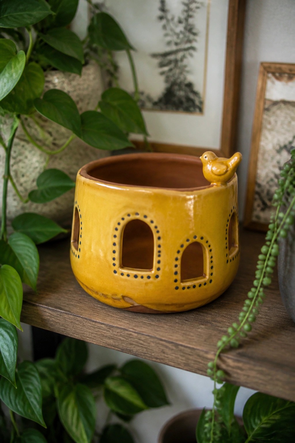 Yellow ceramic house-shaped pot with bird figurine and arched cutouts on wooden shelf