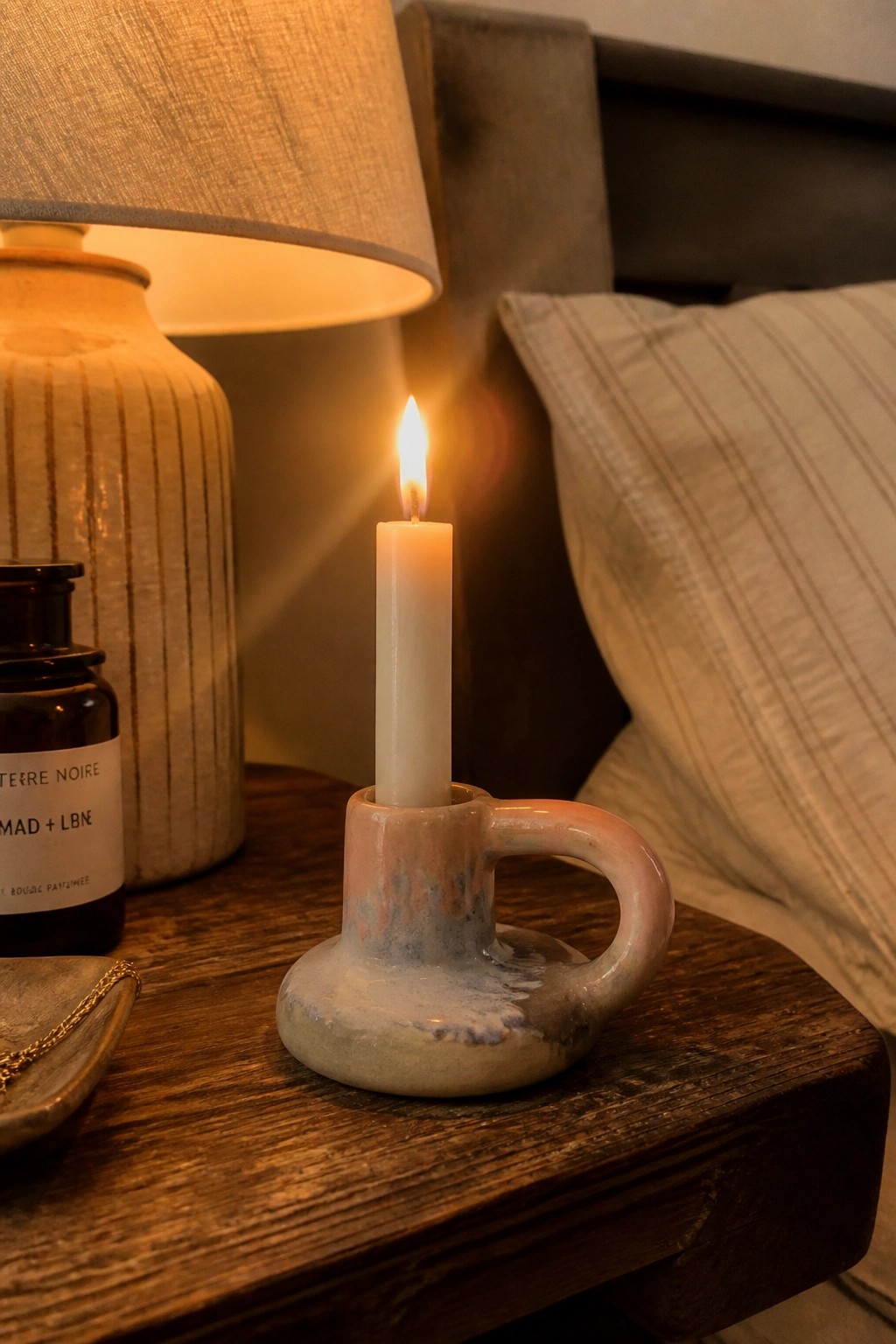 Lit candle in pink-blue ceramic holder on wooden nightstand beside lamp and pillow.