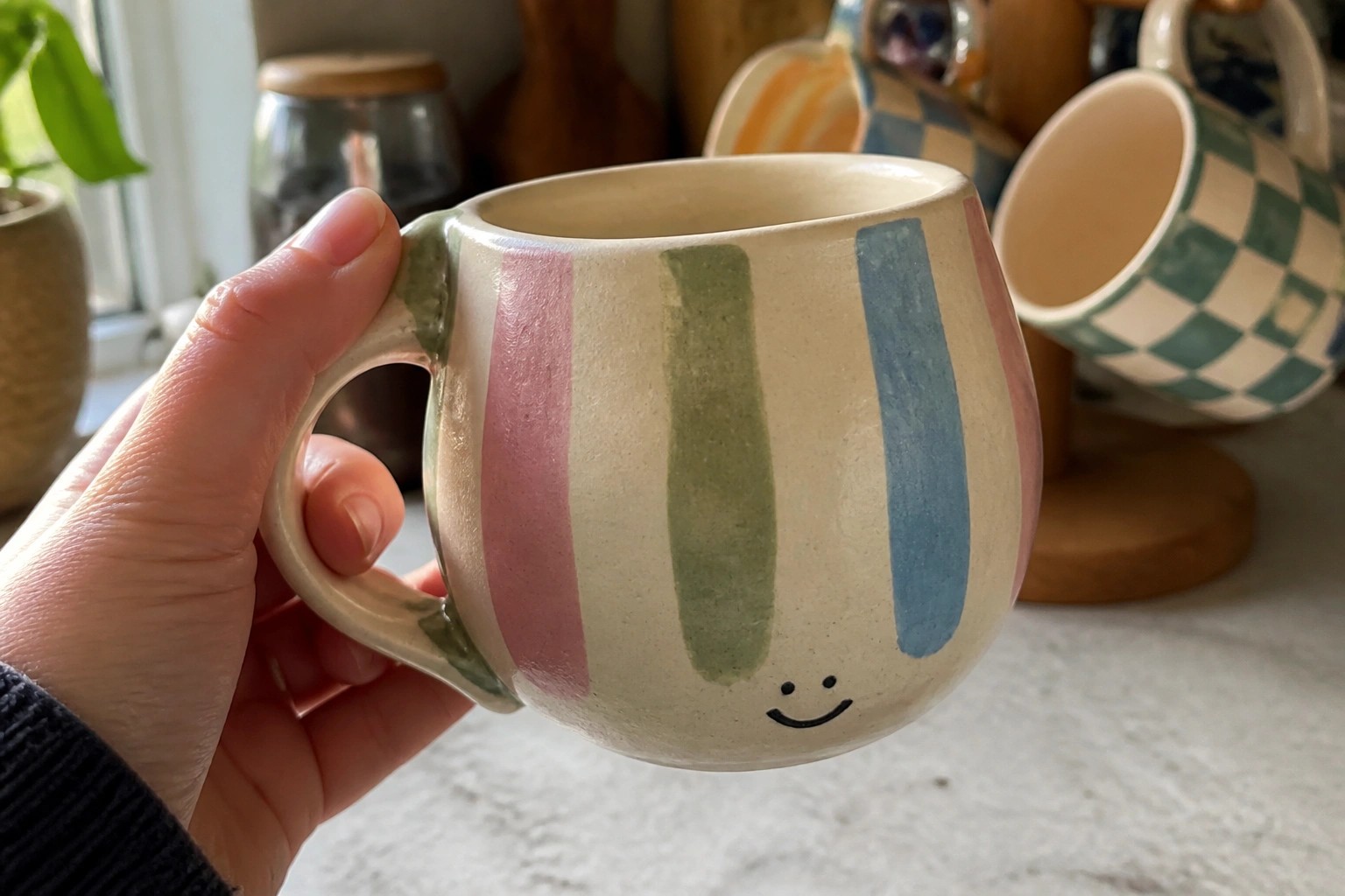 Hand holding a pastel-striped ceramic mug with a smiley face in sunlight.