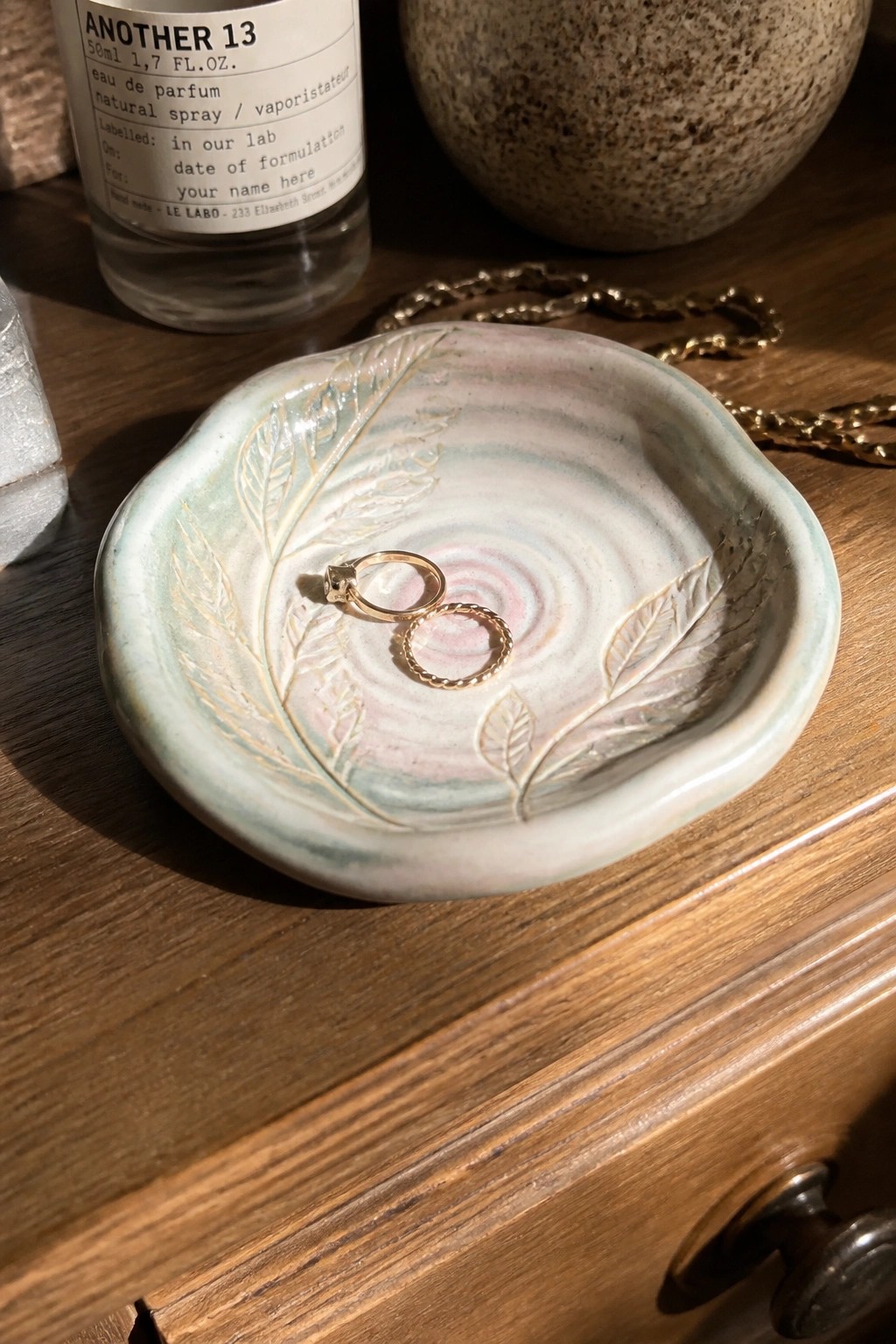 Two gold rings in a leaf-patterned pastel ceramic bowl beside a perfume bottle on wood.