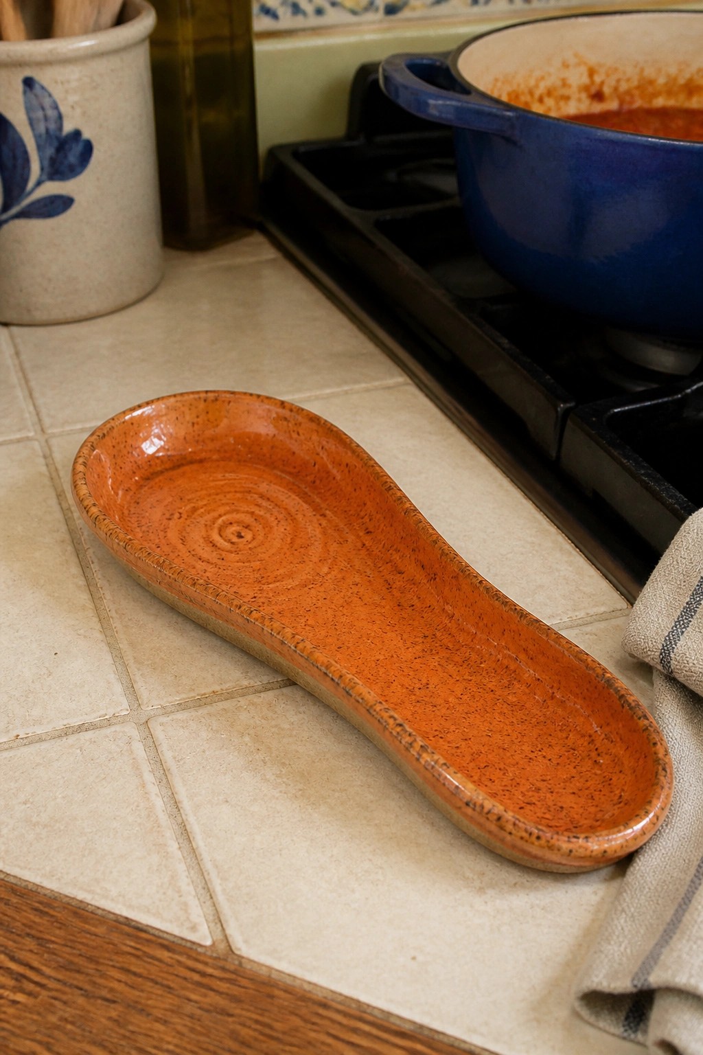 Orange ceramic spoon rest on tiled kitchen counter near blue pot on stove