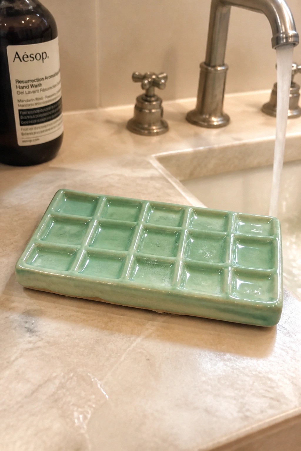 Green ceramic compartmentalized tray on marble sink with running faucet and Aesop bottle.