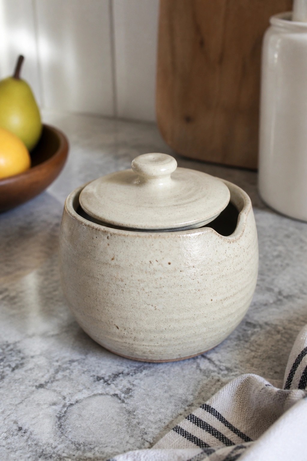 Lidded speckled ceramic pot on marble counter with fruit bowl and striped cloth