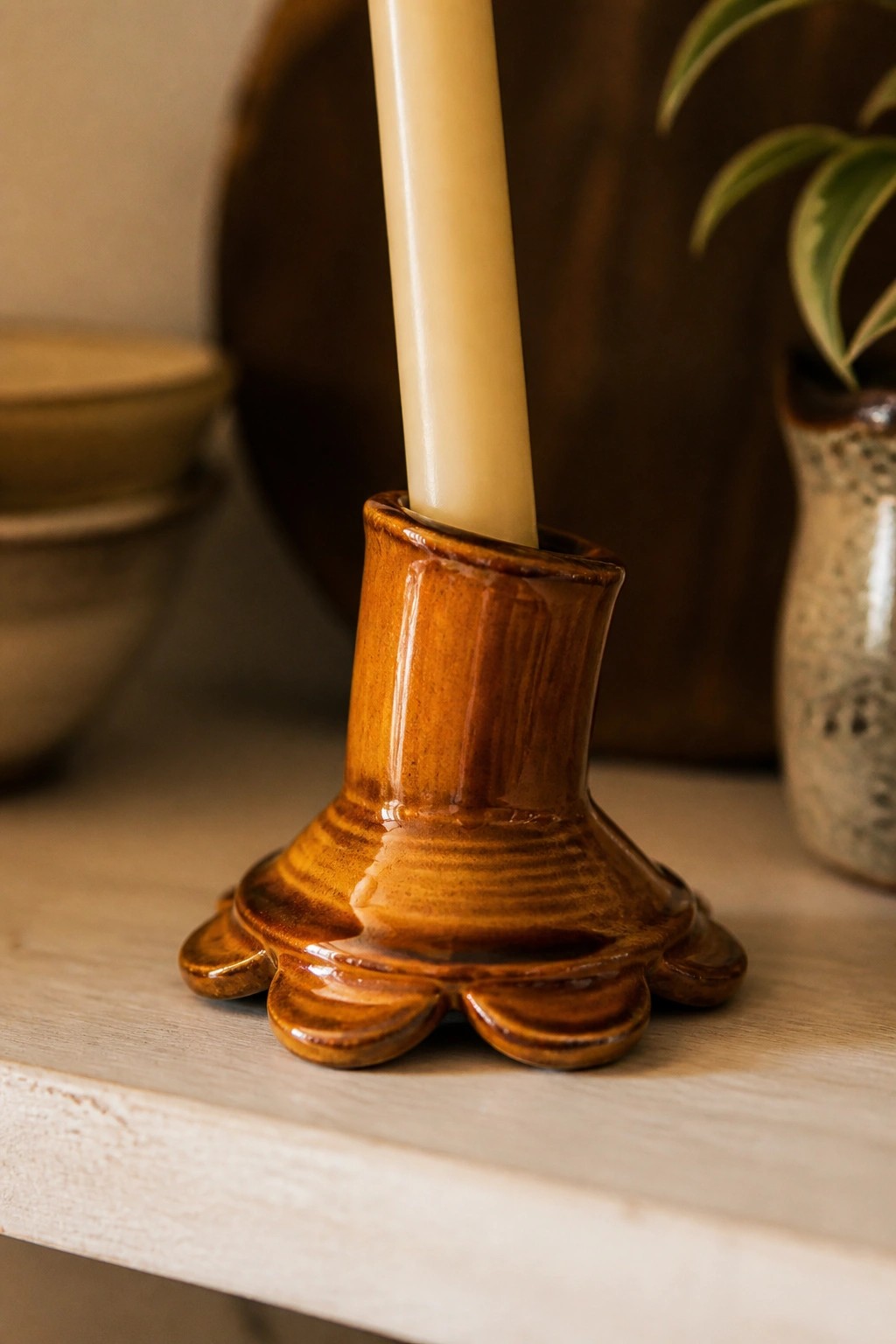 Brown ceramic candlestick with scalloped base holding a tall cream candle on wood.