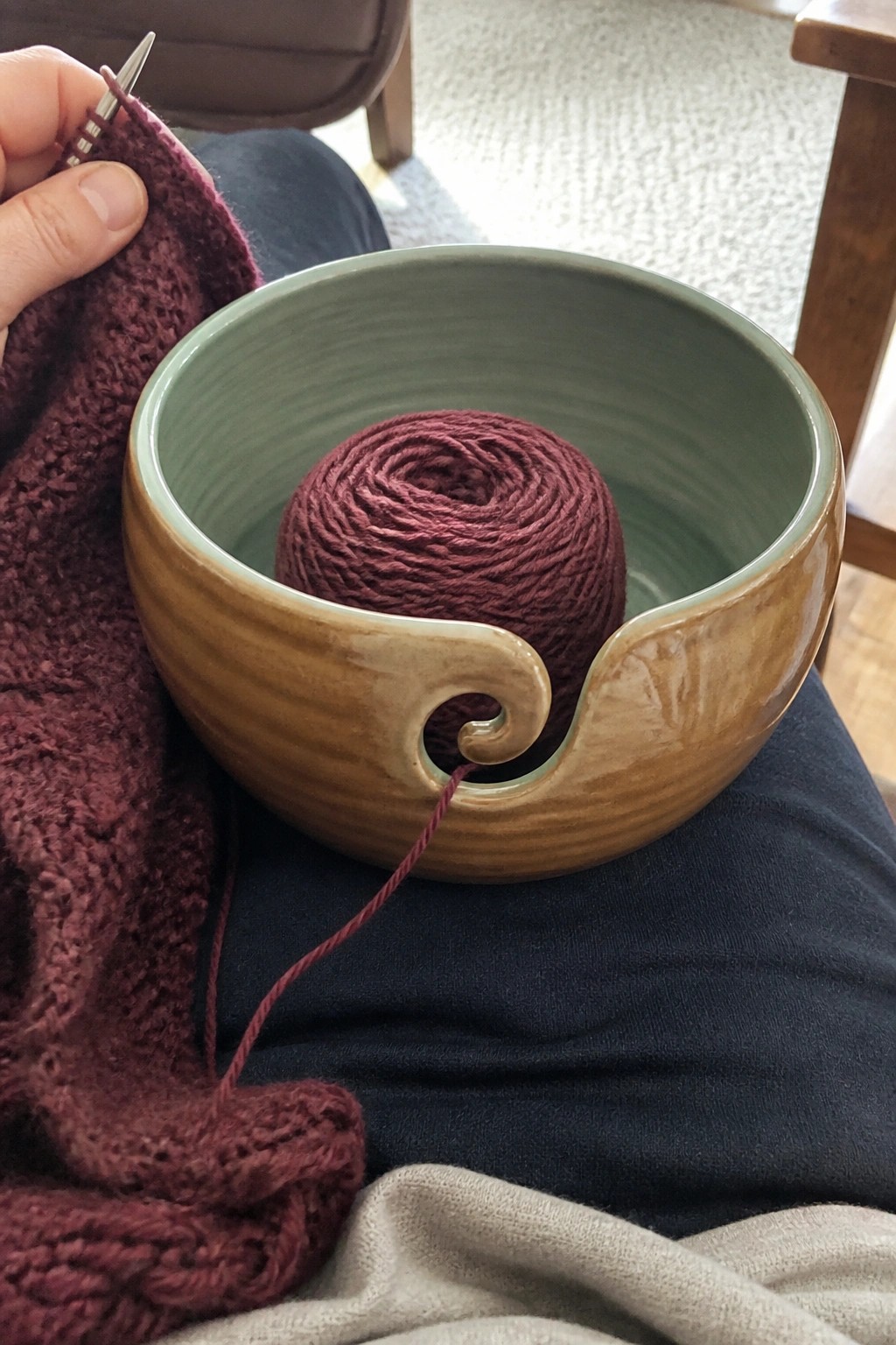 Hands knit maroon fabric beside yarn ball in swirled ceramic bowl on lap