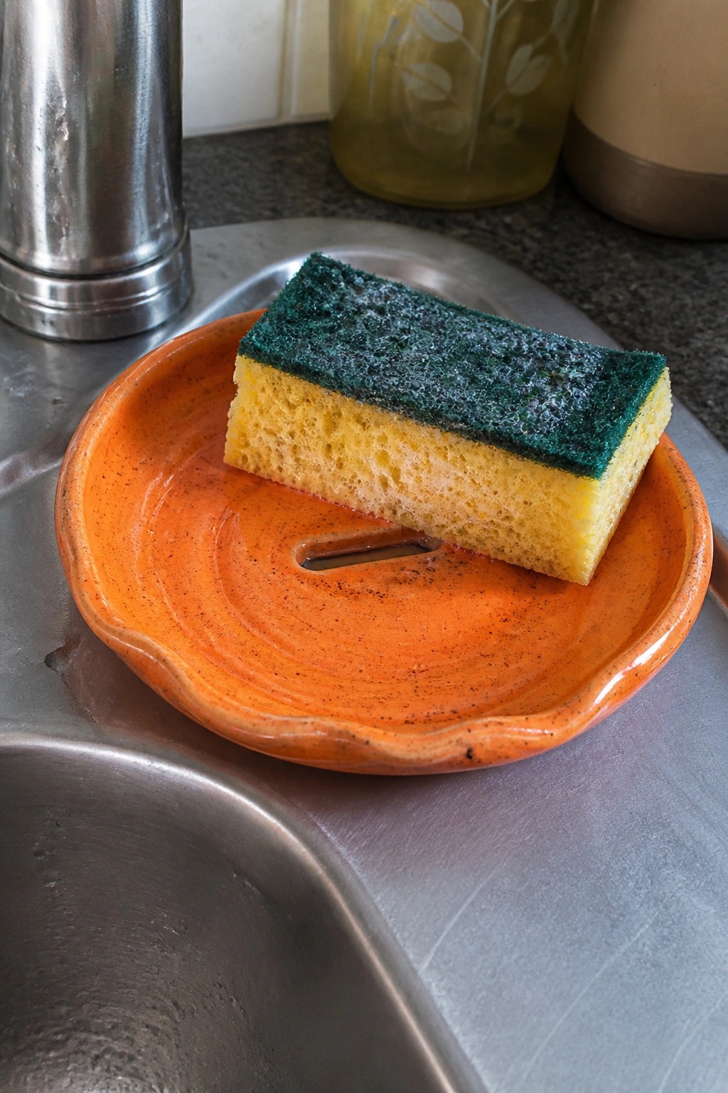 Yellow and green sponge on orange ceramic dish in stainless steel sink.