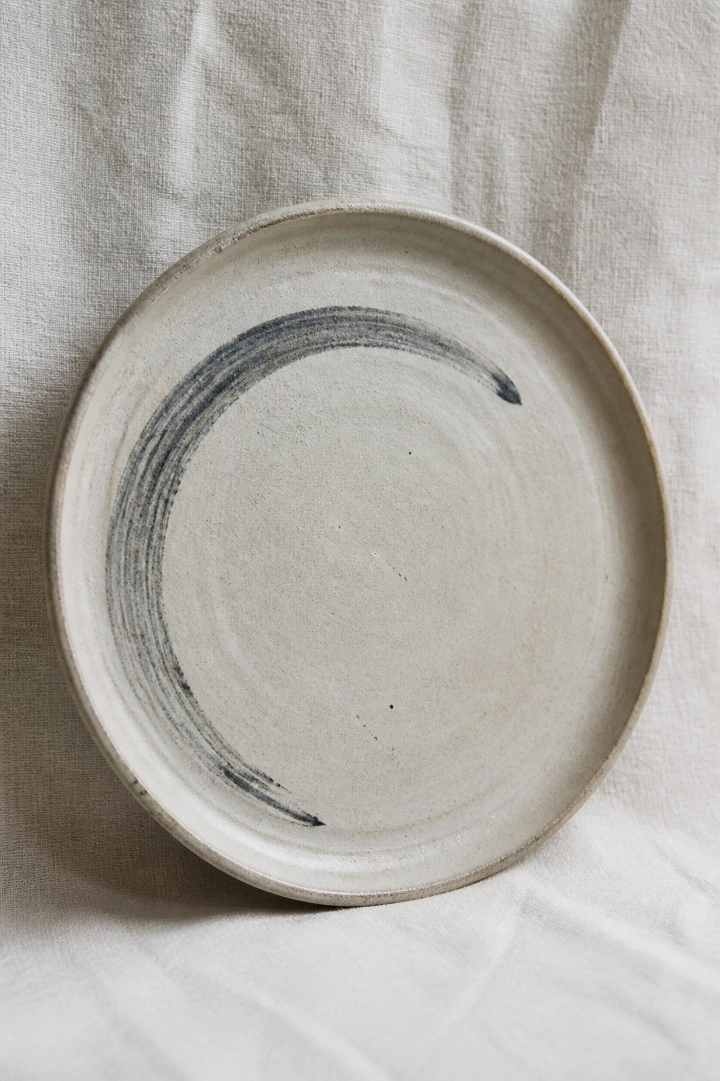 Rustic light ceramic plate with dark curved brushstroke on white linen fabric