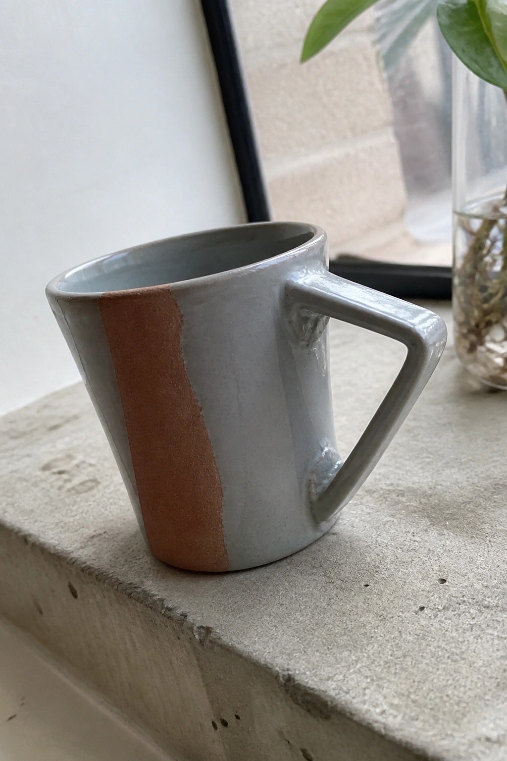 Gray ceramic mug with rust stripe on rough concrete ledge near plant.