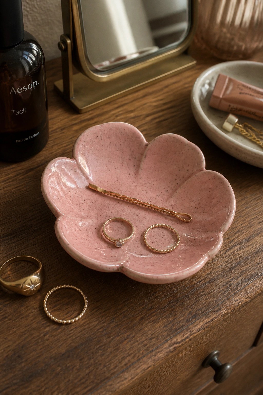 Pink flower-shaped dish holding gold rings and bobby pin on wooden vanity with perfume