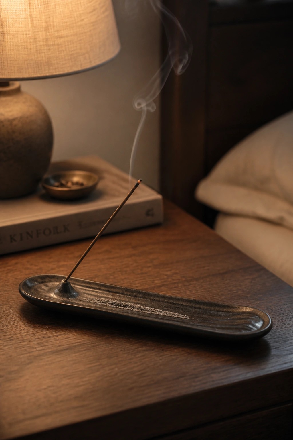 Incense stick burning in dark ceramic tray with rising smoke on wooden nightstand