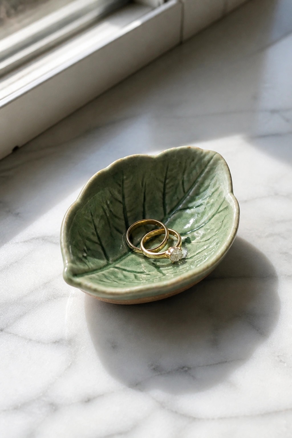Green leaf bowl holding two gold rings on sunlit white marble