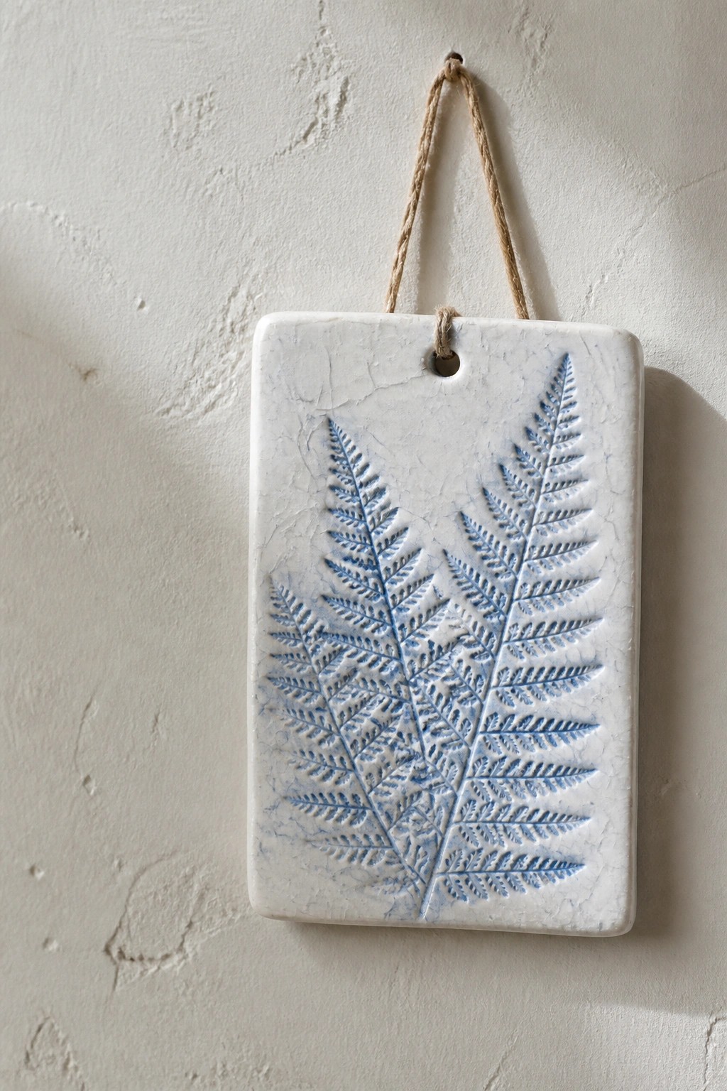 White ceramic tile with blue fern imprints, hanging by twine on textured wall.