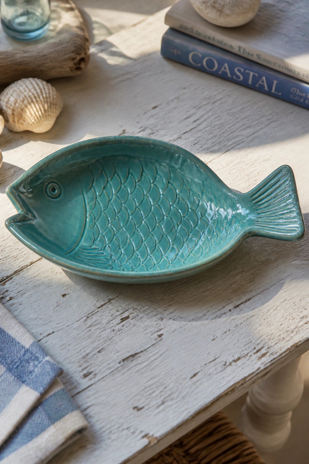 Turquoise ceramic fish-shaped bowl on distressed white wooden table with seashells and books.