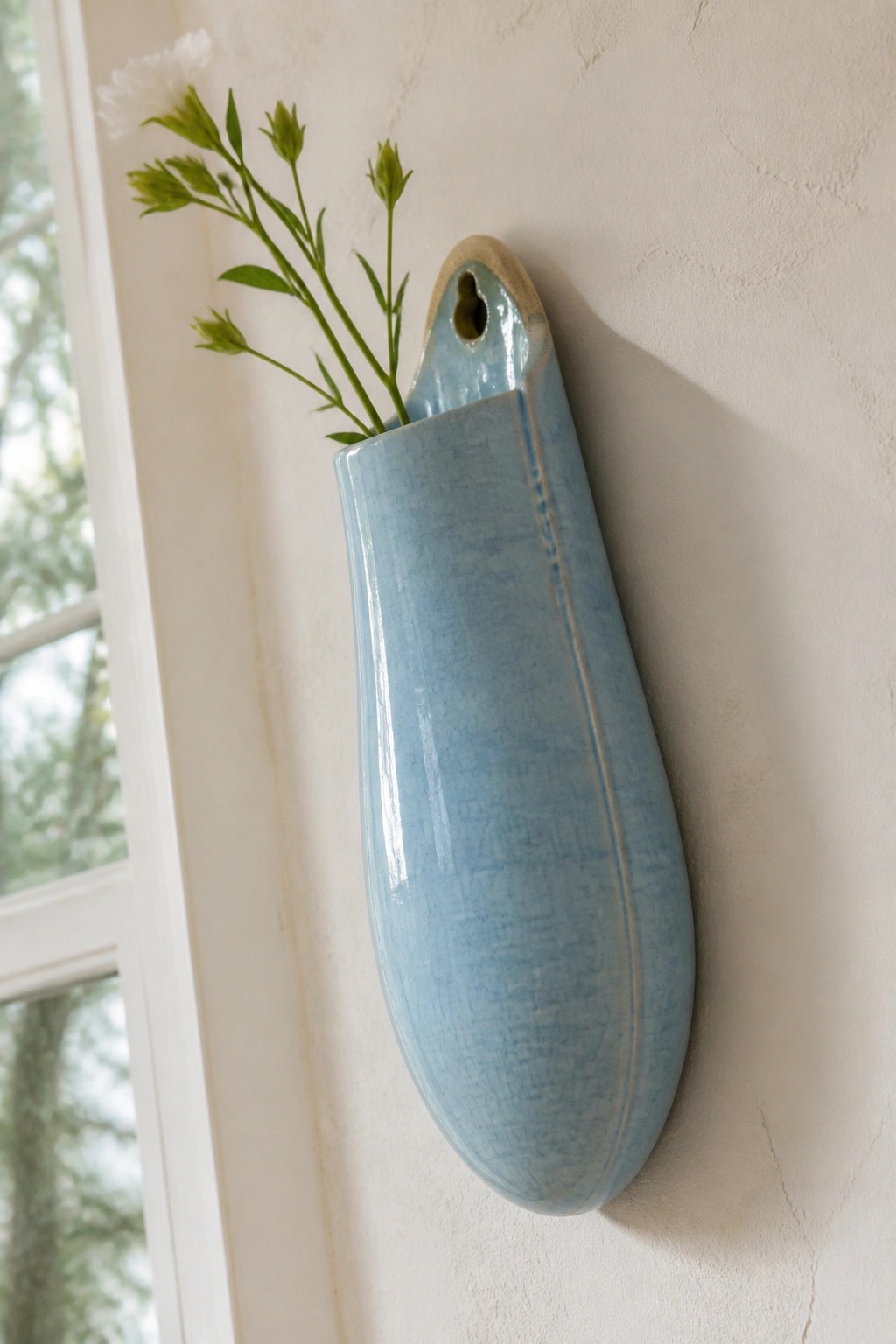 Light blue ceramic wall vase with green stems on textured wall near window