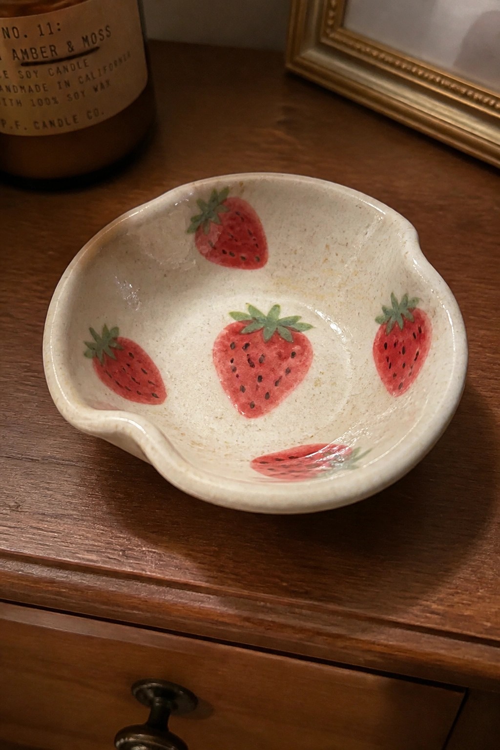 White ceramic bowl with red strawberries on wooden dresser beside candle jar