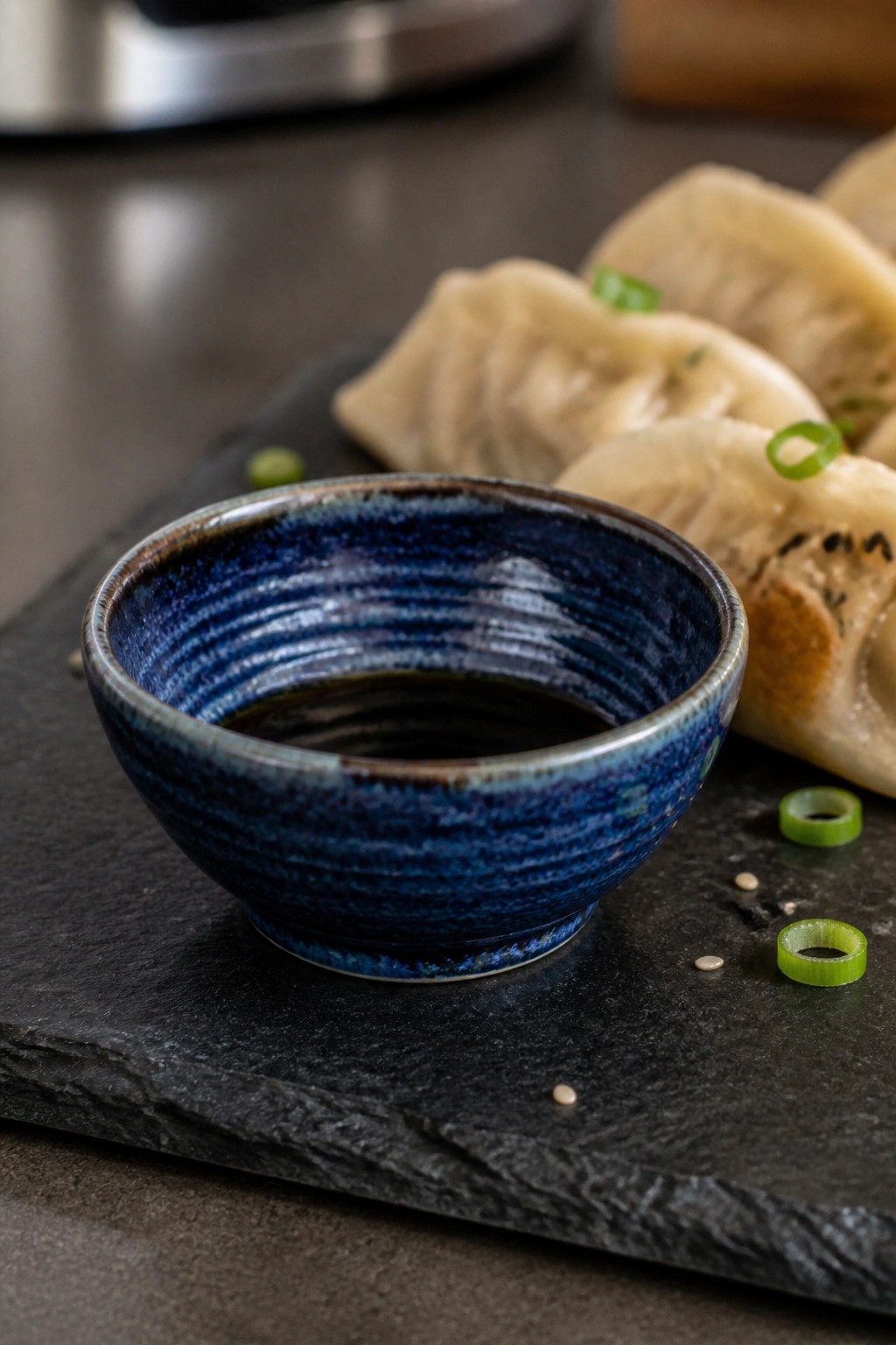 Blue ceramic bowl of soy sauce beside dumplings and green onions on slate